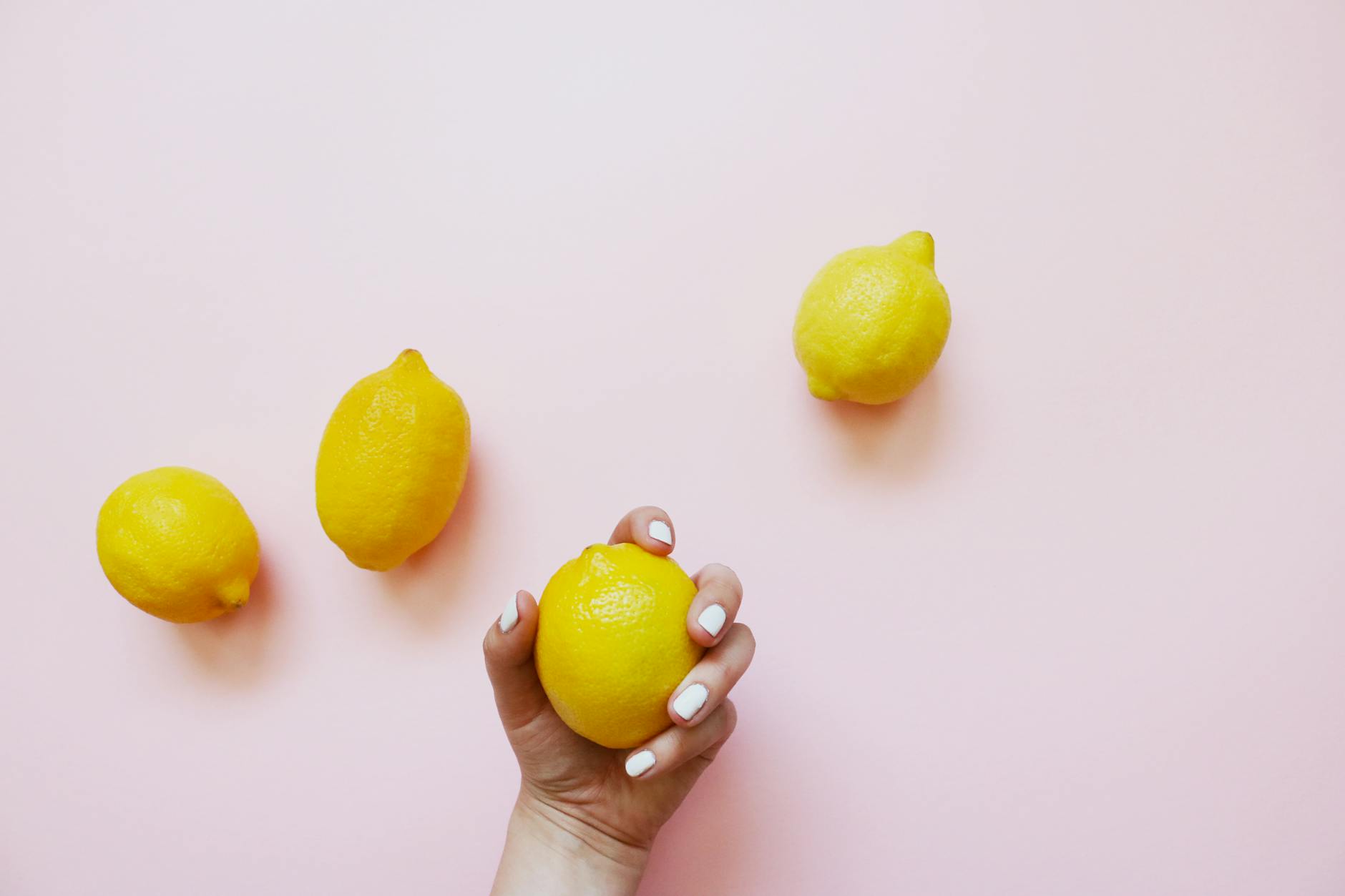 A hand with white nails holding a lemon on a soft pink background surrounded by yellow lemons